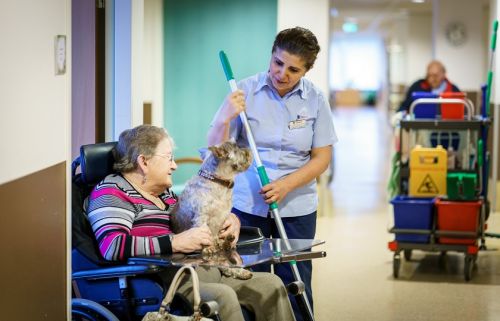 een medewerkster van Gom Zorg staat met een vloerwisser in de hand een praatje te maken met een oudere dame in rolstoel, die een hondje op schoot heeft