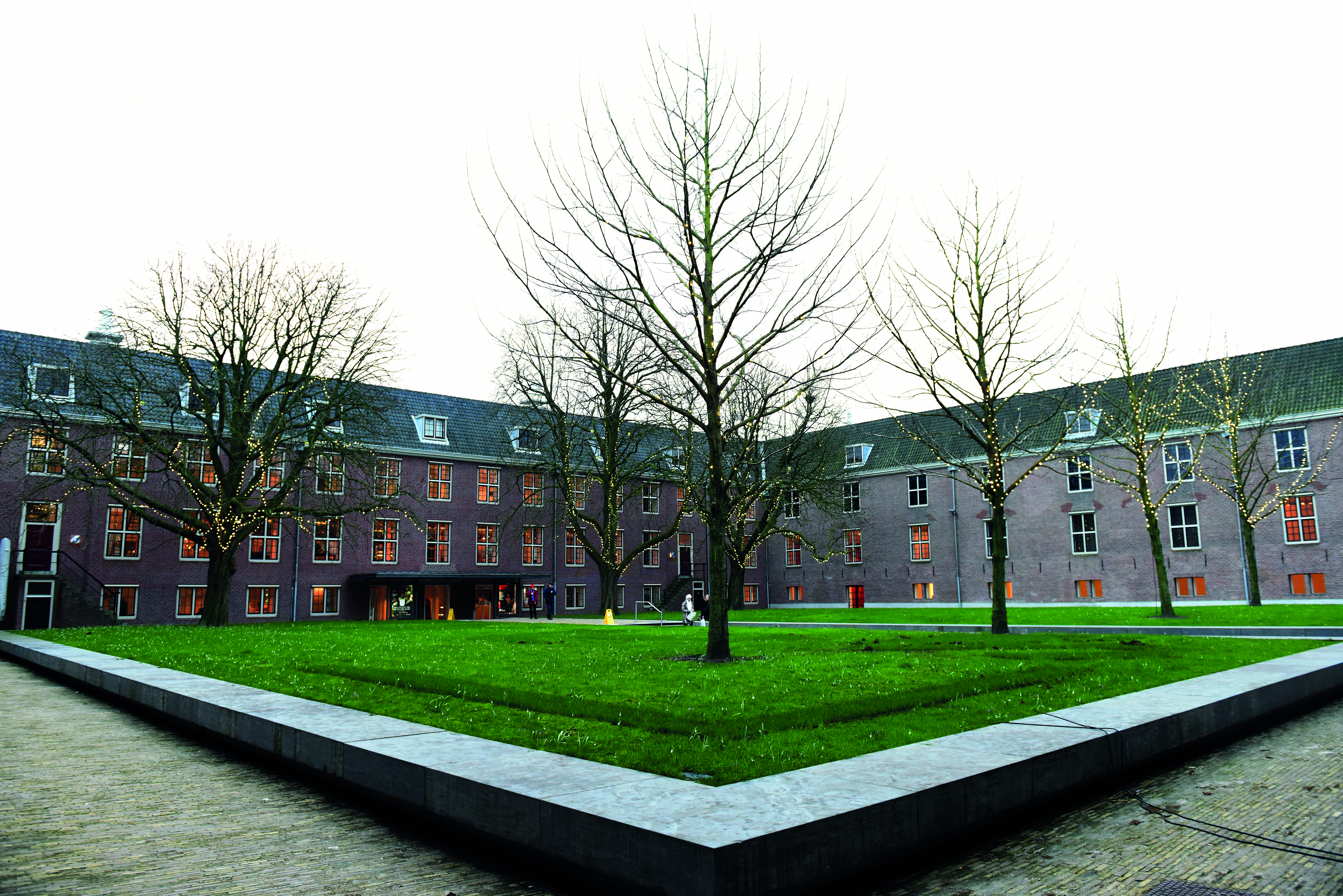 Dit is de binnentuin van de Hermitage in Amsterdam met een groot groen grasveld met verschillende grote bomen.