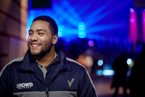 Mannelijke beveiliger werkt op een muziek festival en staat voor de zaal waar het festival plaatsvindt. Hij heeft een uniform aan, lacht naar de camera, maar kijkt opzij.