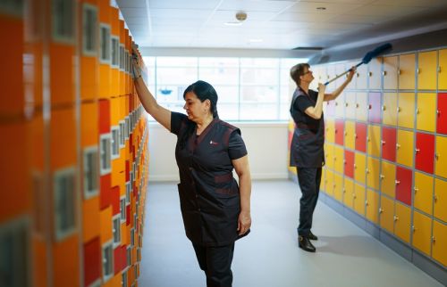 2 medewerksters van Gom maken de geel-oranje-rood gekleurde schoolkluisjes schoon, de een de deurtjes met een doekje, de ander met een stofwisser de bovenkant de de kluisjes