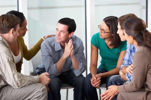 Zes mensen zitten op een stoel en bij elkaar in een kring waarbij zij steun verlenen aan een collega die een heftig verhaal vertelt en hierbij emotioneel is.