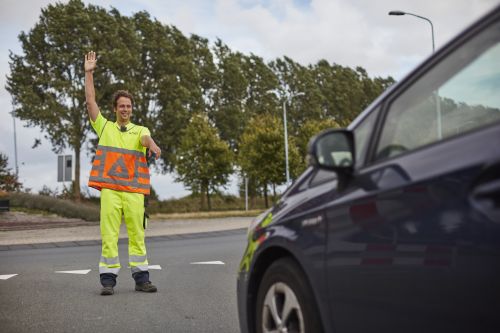 Verkeersregelaar in oranje en geel uniform steekt zijn hand omhoog en houdt een auto tegen.
