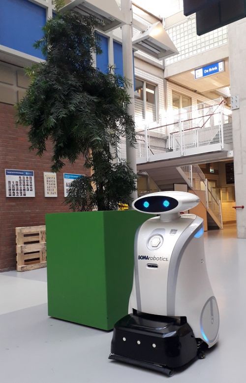 Schoonmaakrobot Ella, een witte robot met digitale ogen, rijdt voorbij een plantenbak