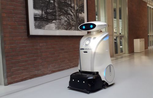 Schoonmaakrobot Ella, een witte robot met digitale ogen, rijdt in een gang voorbij een schilderij