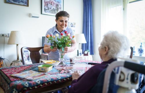 een medewerkster van Gom Zorg schikt de bloemen die op tafel staan bij een oudere dame en kijkt haar lachend aan
