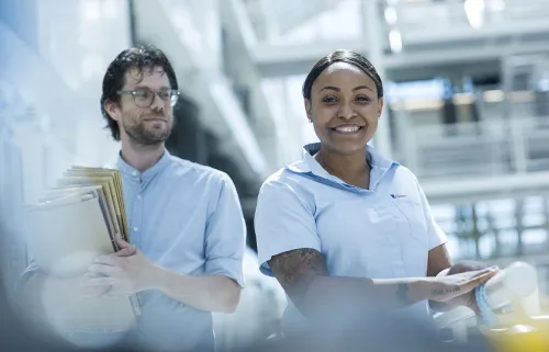een man met mappen in zijn handen loopt een schoonmaakmedewerkster voorbij die in de camera kijkt en een doekje over de reling haalt.