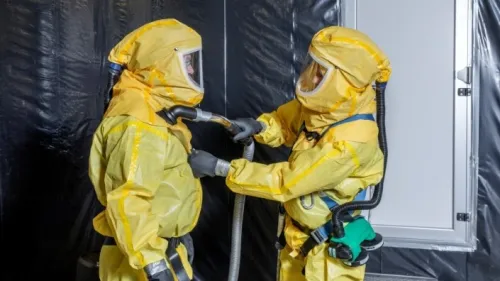 Specialist van Gom in gele veiligheidsuitrusting verwijdert fijnstof met een industriële stofzuiger bij een ventilatie-installatie in een fabriekshal.