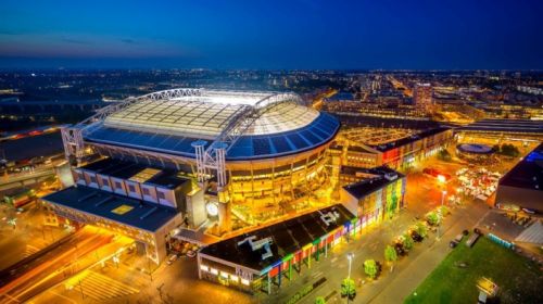 De kijk van bovenaf op de Johan Cruijff Arena in Amsterdam in de avond waarbij zowel de arena als het treinstation is verlicht.