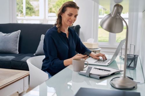 Een vrouw kijkt op haar telefoon terwijl ze achter haar laptop zit te werken