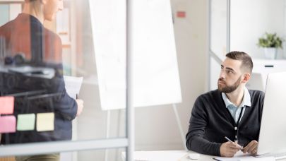 Twee collega's in gesprek. Een van de collega's zit achter zijn bureau met daarop een beeldscherm maar kijkt zijn collega aan die naast hem staat Twee collega's in gesprek. Een van de collega's zit achter zijn bureau met daarop een beeldscherm maar kijkt zijn collega aan die naast hem staat