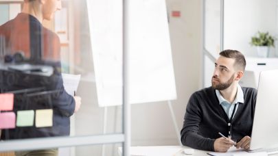 Twee collega's in gesprek. Een van de collega's zit achter zijn bureau met daarop een beeldscherm maar kijkt zijn collega aan die naast hem staat