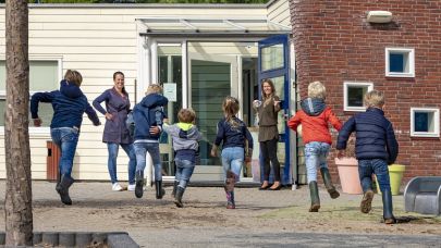 een groepje basisschoolkinderen rent vol enthousiasme richting de deur van het schoolgebouw waar de juf en de schoonmaakster van Gom ze staan op te wachten