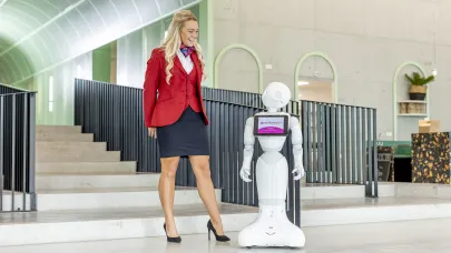 Gastvrouw in uniform staat naar robot in centrale hal van een kantoorpand.