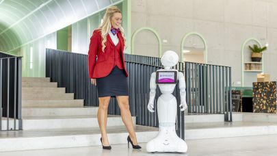 Gastvrouw  in uniform staat naar robot in centrale hal van een kantoorpand.