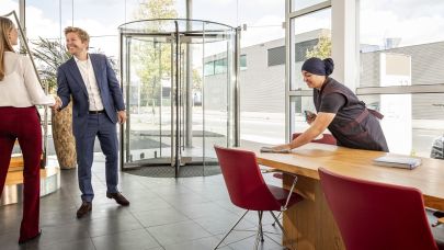een bezoeker komt een bedrijfspand binnen en wordt verwelkomd door een medewerker en ze schudden de hand, ondertussen is een schoonmaakmedewerkster van Gom de leestafel aan het schoonmaken.