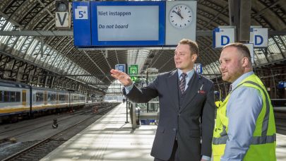 Mannelijke beveiliger in uniform legt aan mannelijke beveiliger met een veiligheidshesje uit wat de situatie is. Zij staan samen op het perron van een treinstation.