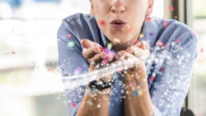 Een jonge vrouw kijkt in de camera en blaast gekleurde confetti uit haar handen.