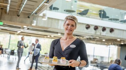 Vrouwelijke Embracemedewerkster heeft een dienblad vast met daarop verschillende taartjes. Vrouwelijke Embracemedewerkster heeft een dienblad vast met daarop verschillende taartjes.