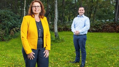 een foto van Astrid Jeurissen, hoofd facilitaire services Sherpa en Rick Werring, rayonleider Gom Zorg, buiten op een grasveld omringd door bomen en bossages