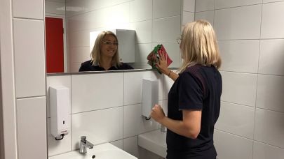 Schoonmaakmedewerkster Beata Gorzelanczyk kijkt je via de spiegel in de toiletruimte aan, terwijl ze de spiegel schoonmaakt met een blauw glazendoekje