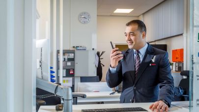 Mannelijke beveiliger in uniform praat via de portofoon met een collega beveiliger op locatie en kijkt daarnaast ook gelijk op zijn computerscherm achter zijn bureau.