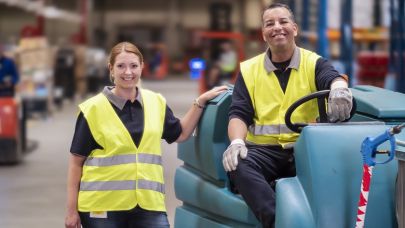 Twee schoonmaakmedewerkers van Gom Logistiek kijken lachend in de camera, 1 hiervan zit op een schrobzuigmachine, de ander staat ernaast