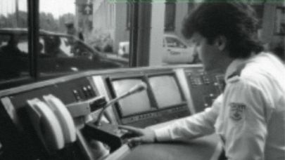 Zwart-wit foto met mannelijke beveiliger achter verouderde computerschermen sprekend in een microfoon. 