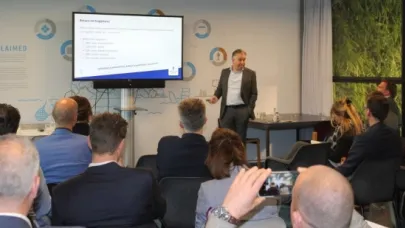 Spreker geeft een presentatie aan een groep aanwezigen, met een projectiescherm waarop een dia met tekst wordt getoond.