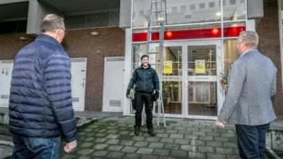Monteur staat onderaan een ladder bij de ingang van een gebouw, terwijl twee mannen toekijken.
