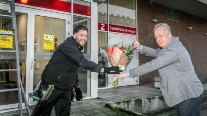 Man in grijs pak overhandigt een bos bloemen aan een monteur in werkkleding voor de ingang van een gebouw.