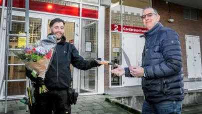 Iemand ontvangt bloemen en een envelop uit handen van een collega voor de ingang van een gebouw.