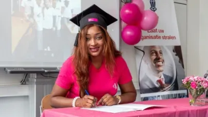 Vrouw in roze shirt en afstudeerhoed ondertekent een document tijdens een ceremonie, met ballonnen en bloemen op de achtergrond.