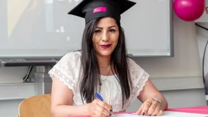Vrouw met afstudeerhoed tekent een document tijdens een diploma-uitreiking, zittend aan een tafel met roze tafelkleed.