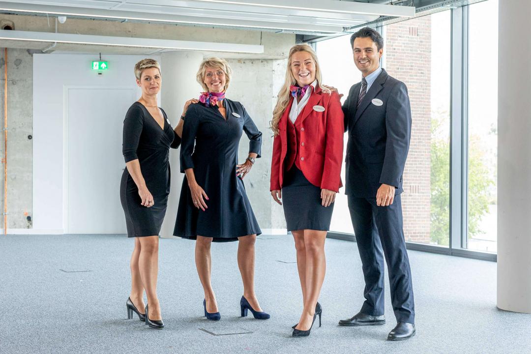 Drie vrouwelijke receptionisten en een gastheer staan geposeerd voor de camera in hun uniformen.