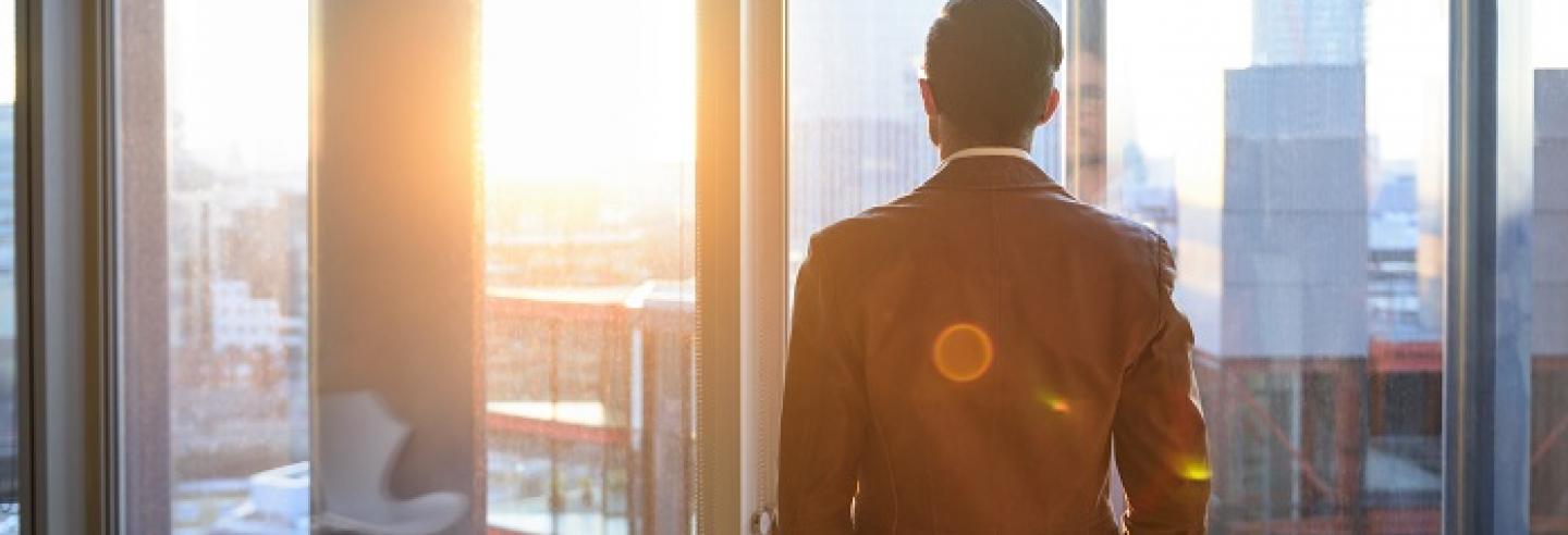 Man in zakelijke kleding kijkt uit het raam van zijn kantoor.