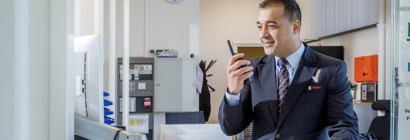 Mannelijke beveiliger in uniform praat via de portofoon met een collega beveiliger op locatie en kijkt daarnaast ook gelijk op zijn computerscherm achter zijn bureau.