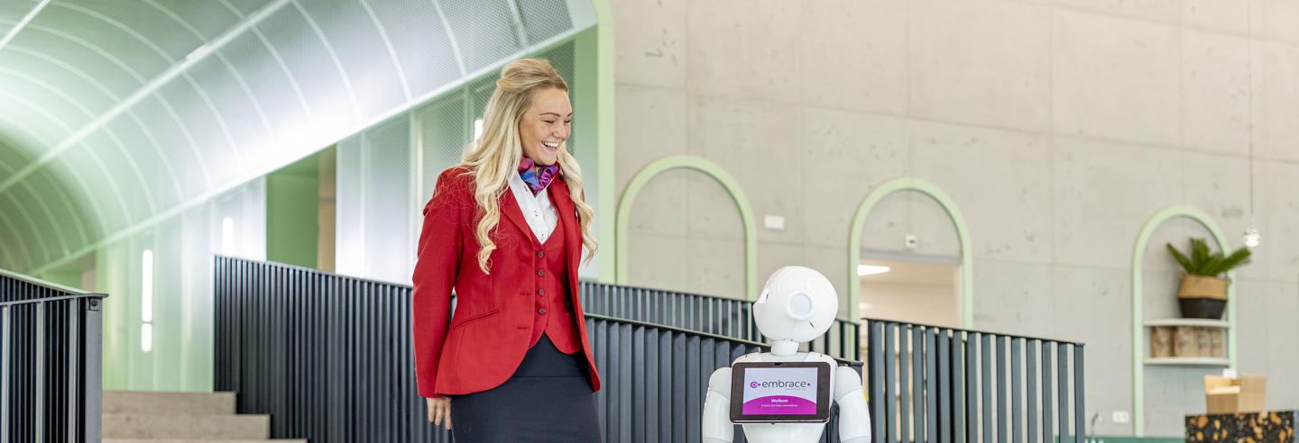 Gastvrouw  in uniform staat naar robot in centrale hal van een kantoorpand.