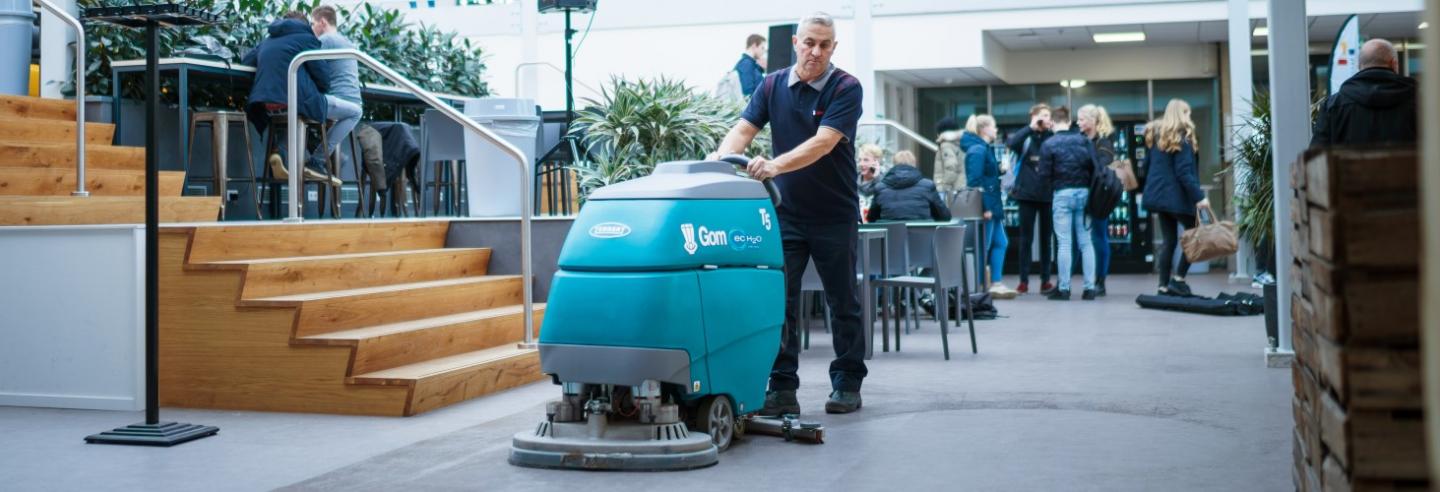 een medewerker van Gom is met een schrobzuigmachine de vloer aan het reinigen in een schoolpand