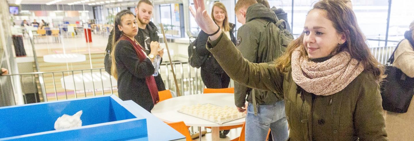 een studente gooit met een boogje een propje papier in de trash roulette, de interactieve afvalbak van Gom Onderwijs, op de achtergrond kijken studenten mee