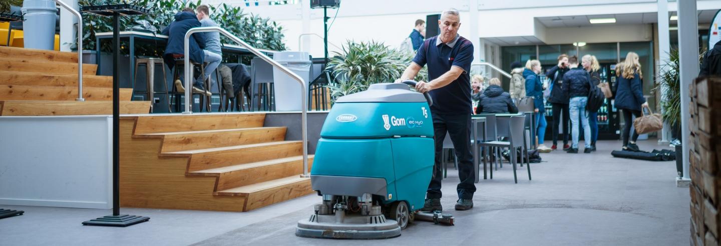 een medewerker van Gom Onderwijs maakt met een schrobzuigmachine de vloer in de schoolhal schoon, op de achtergrond staat een groepje leerlingen