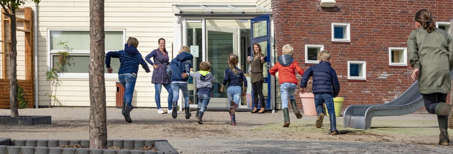 een groepje basisschool-kinderen rennen naar de deur van de school waar zowel de juf als de schoonmaakster van Gom ze staat op te wachten