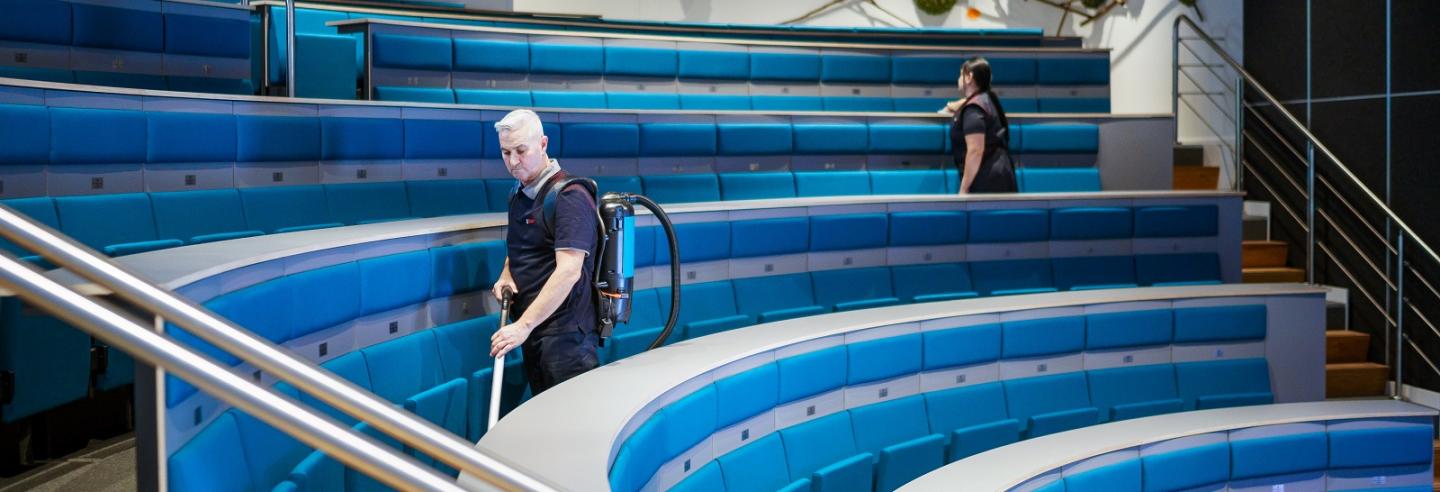 2 schoonmaakmedewerkers maken schoon in een auditorium van een mbo-school, de een stofzuigt, de ander neemt de stoelen af.