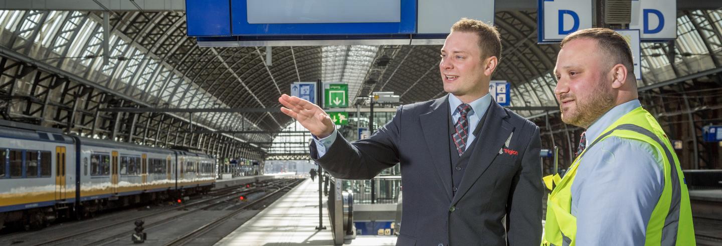 Mannelijke beveiliger in uniform legt aan mannelijke beveiliger met een veiligheidshesje uit wat de situatie is. Zij staan samen op het perron van een treinstation.