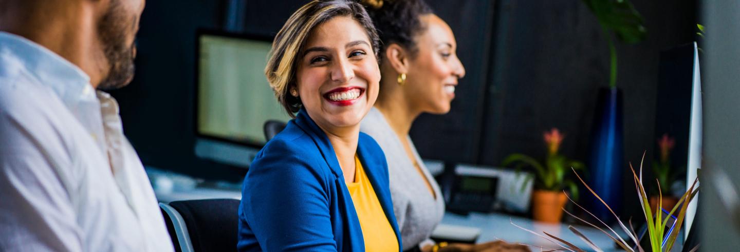 Een man en twee vrouwen zitten naast elkaar aan een bureau achter de computer, de focus ligt op een vrouw met een blauw jasje en een geel shirt.