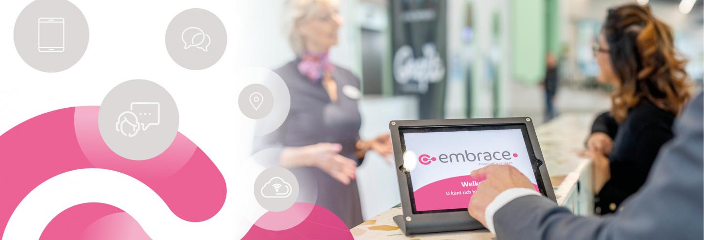 Je ziet de hand van een bezoeker die zich aanmeldt op een tablet met daarop het logo van Embrace. Achter de tablet zie je een Embrace-medewerkster in gesprek met een bezoeker.