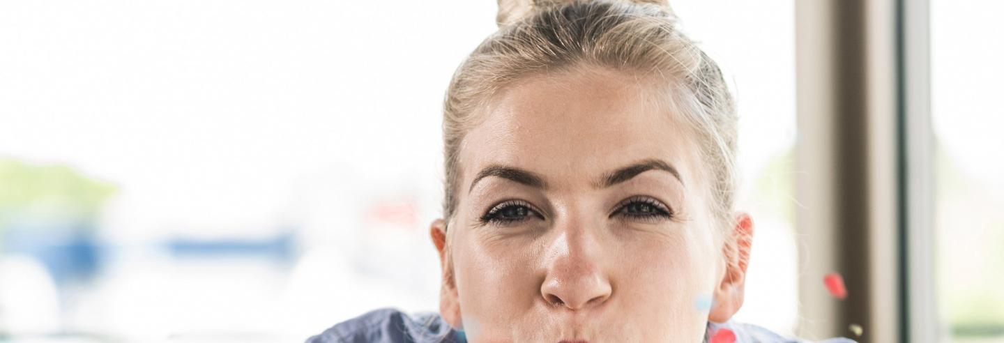 Een jonge vrouw kijkt in de camera en blaast gekleurde confetti uit haar handen.