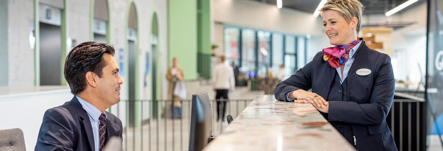 Mannelijke receptionist in uniform zit achter de balie en praat met een gastvrouw in uniform aan de andere kant van de balie.