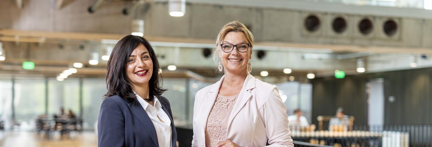 Twee vrouwen in zakelijke kleding staan geposeerd voor de camera in een lichte ruimte.