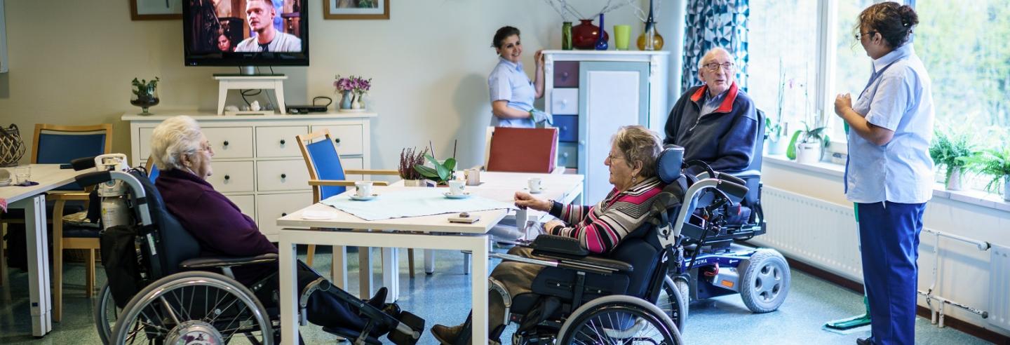 drie ouderen in een rolstoel zitten om een tafel, 2 medewerkers van Gom Zorg doen de schoonmaak