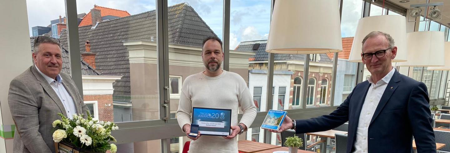 Drie mannen staan geposeerd voor de camera waarbij de medewerker een award in zijn handen heeft en de andere twee mannen de bloemen en de voucher.